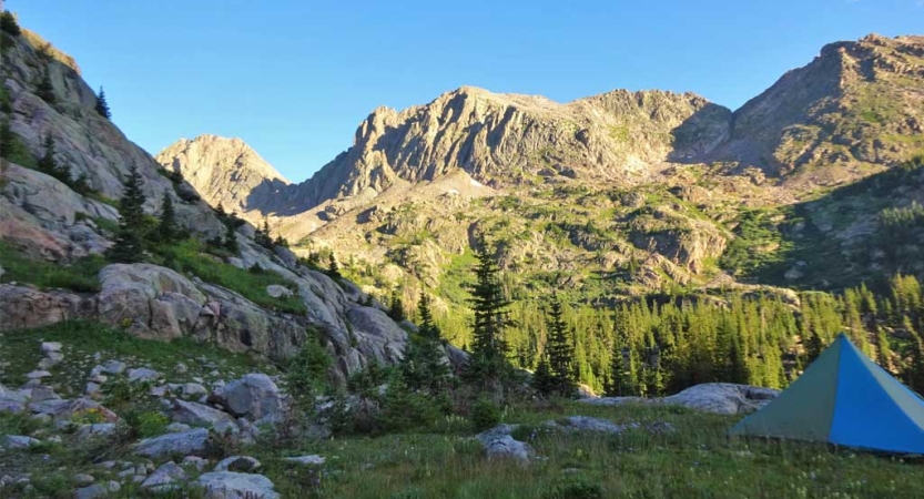 A tent rests in a green meadow amid tall rocky mountains